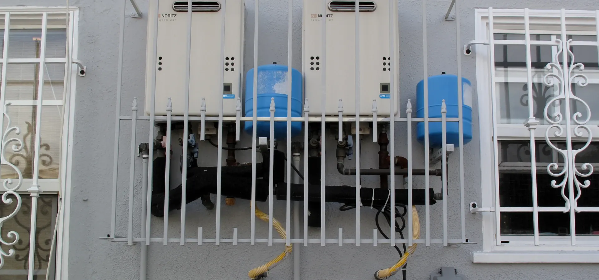Two wall-mounted tankless water heaters with expansion tanks behind a barred window