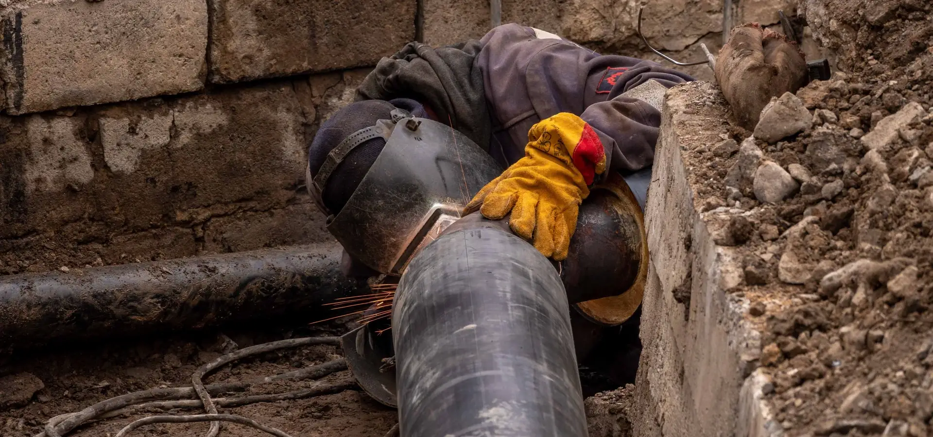 Worker in a welding helmet and yellow gloves joining a large pipe in an excavation